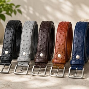 Puede incluir: Cinco cinturones con un patrón texturizado estilo avestruz, cada uno con una hebilla de metal plateado y un botón a presión blanco cerca de la base. Los cinturones están dispuestos en fila y vienen en colores negro, gris claro, marrón oscuro, tostado y azul marino.