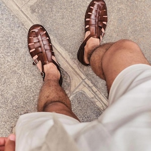 May include: A pair of dark brown leather fisherman sandals featuring a closed-toe design with woven straps, a buckled ankle strap, and a sturdy sole. The sandals are worn with light-colored shorts, showcasing the cage-like upper construction.