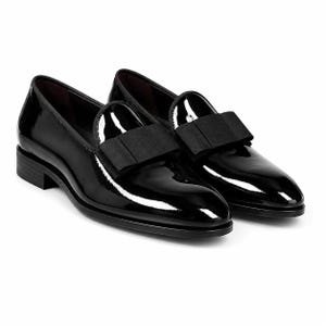 Op de afbeelding: Een paar zwarte lakleren loafers met een glanzende afwerking en een gestructureerde zwarte stoffen strik op de wreef. Deze instapschoenen hebben een ronde neus, een lage blokhak en een donkere voering, wat zorgt voor een strakke uitstraling.