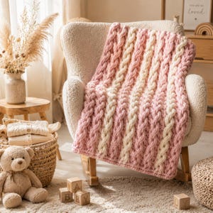 Può includere: Una coperta in maglia grossa con strisce verticali alternate di filato rosa tenue e crema. La coperta presenta una trama a trecce prominente su tutta la superficie, con spessi motivi ritorti simili a corde che corrono in verticale.