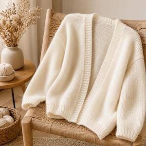 May include: A cream-colored open-front cardigan sweater featuring a soft knit texture, long sleeves with ribbed cuffs, and a ribbed trim along the front opening. The garment is displayed draped over a woven chair, showcasing its simple design.
