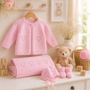 Può includere: Un set per neonati in maglia rosa chiaro composto da un cardigan a maniche lunghe con un bottone, una coperta abbinata, un berretto con nastri e scarpine. Ogni pezzo presenta un delicato motivo traforato a forma di cuore nel filato.