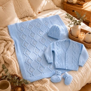 Puede incluir: Un conjunto de punto para bebé en color azul claro que incluye una manta con patrón de rombos, una chaqueta de manga larga con botones y detalles de trenzas, un gorro a juego y un par de patucos, todo con una textura suave.