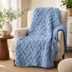 May include: A light blue knitted throw blanket featuring a prominent basketweave pattern with thick, textured yarn. The blanket is draped over a neutral-colored upholstered armchair, showcasing the intricate crisscross design of the knit.