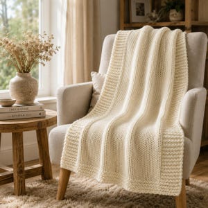 Può includere: Una coperta in maglia color crema con un motivo a coste testurizzato e un ampio bordo a legaccio. La coperta è drappeggiata su una poltrona imbottita di colore chiaro, mettendo in risalto la sua lavorazione a maglia spessa e il tono neutro.