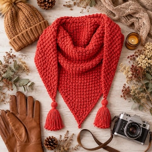 Puede incluir: Una bufanda triangular de punto en color rojo vibrante con un patrón texturizado y dos borlas de hilo a juego en las esquinas inferiores. La bufanda se muestra junto a un gorro de punto beige, guantes de cuero marrón y una cámara vintage.