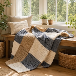 May include: A chunky knit throw blanket featuring a large checkered pattern in alternating blocks of cream, tan, and slate gray. The blanket has a textured, woven appearance and is draped over a wooden bench with a matching cream knit pillow.