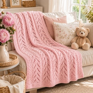 May include: A soft, light pink knitted throw blanket featuring a vertical chevron and openwork stitch pattern. The blanket is draped over a sofa, showcasing its textured yarn construction and finished edges, providing a cozy and tactile appearance.