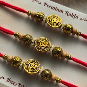 May include: Three red thread bracelets, each featuring a central gold-toned circular charm with an Om symbol, flanked by ornate gold-toned beads, small white seed beads, and light wooden beads, displayed on a card labeled Premium Rakhi.