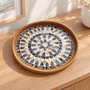 May include: A round serving tray with a woven light brown rattan rim and a flat base featuring a decorative mosaic pattern. The mosaic consists of teardrop-shaped pieces in shades of dark gray, cream, and tan arranged in concentric circular rings.