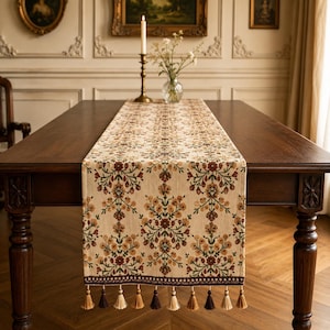 Peut inclure: Un chemin de table rectangulaire avec une base crème ornée d'un motif floral répétitif dans des tons de brun, d'orange et de vert. Le chemin de table est bordé d'une bordure brun foncé et d'une série de pompons suspendus aux couleurs alternées.