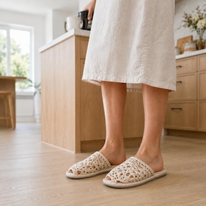 May include: A pair of off-white crochet slippers featuring an intricate openwork pattern, a rounded toe, and a flat sole. The slippers are crafted from a light-colored yarn and provide a breathable, textured design for indoor wear.