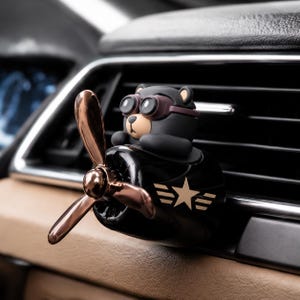 Può includere: Un deodorante per auto nero a forma di motore di aereo vintage con un orso pilota che indossa occhiali protettivi marroni. La parte anteriore presenta un'elica color rame metallizzato e un emblema a stella dorata con ali orizzontali.