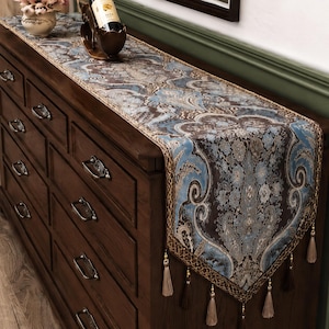 May include: A long table runner featuring a dark brown and light blue damask pattern with intricate floral motifs. The runner has a gold-toned border and is finished with multiple hanging tassels along the edges, draped over a wooden dresser.