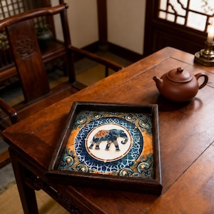 May include: A square wooden tray featuring a central circular medallion with a blue elephant illustration. The tray has a dark brown frame and a decorative border with blue, gold, and orange patterns surrounding the elephant motif.