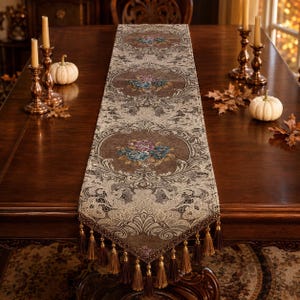 Puede incluir: Un camino de mesa rectangular largo con una base de tela marrón, intrincados patrones de encaje color crema y medallones florales centrales en azul, morado y dorado. Los extremos están rematados con borlas decorativas y flecos marrones.