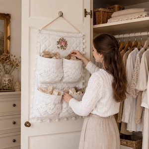 Könnte beinhalten: Ein weißer, gesteppter Wand-Organizer aus Stoff zum Aufhängen an einer Tür, mit vier Taschen mit Spitzenbesatz und einer floralen Stickerei oben. Der Organizer wird an einer Kordel aufgehängt und ist mit kleinen weißen Bommel-Fransen verziert.