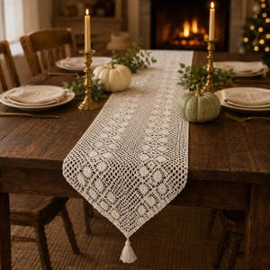 May include: A cream-colored crocheted table runner with a repeating floral pattern and a tassel at the end. The runner is placed on a dark wooden table with white and green pumpkins, two brass candlesticks with lit candles, and place settings.