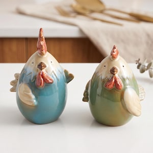 Può includere: Due statuette di pollo in ceramica con finiture lucide. Il pollo a sinistra è blu con il petto color crema e cresta e bargigli rossi. Il pollo a destra è verde con il petto color crema e cresta e bargigli rossi. Entrambi hanno piccole ali.
