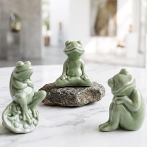 Könnte beinhalten: Drei hellgrüne Keramikfroschfiguren mit glänzender Oberfläche. Ein Frosch sitzt in meditativer Haltung auf einem dunkelgrauen, strukturierten Stein, während die anderen beiden Frösche seitlich davon positioniert sind.