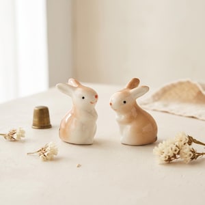May include: Two small ceramic rabbit figurines with a glossy white and light tan finish, featuring painted black eyes and orange noses. A metallic thimble and small dried white flowers are positioned near the two sitting bunny sculptures.