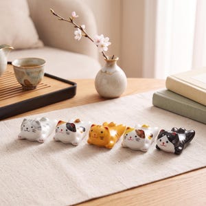 Puede incluir: Un conjunto de cinco reposa palillos de cerámica con forma de gatos tumbados, cada uno con patrones de pelaje distintos que incluyen gris atigrado, calicó, naranja atigrado, blanco con manchas marrones y blanco y negro, con rasgos faciales pintados.