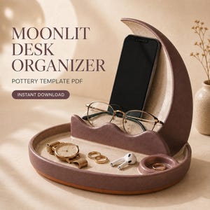 May include: A crescent moon-shaped pottery desk organizer in a muted mauve and speckled cream finish. The organizer features a vertical slot for a smartphone, a horizontal tray for accessories, and a small circular indentation for rings.