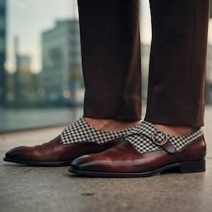 Puede incluir: Un par de zapatos de cuero marrón con hebilla y un panel de tela con estampado de pata de gallo en blanco y negro sobre el empeine. Los zapatos tienen punta redondeada, un cierre de hebilla metálica y se usan con pantalones marrón oscuro.
