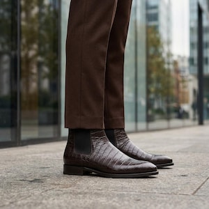 Puede incluir: Un par de botines Chelsea de color marrón oscuro con un patrón texturizado de imitación de cocodrilo, paneles laterales elásticos negros y tacón bajo, combinados con pantalones marrón oscuro que llegan hasta el tobillo.