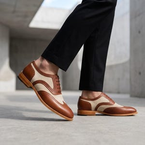 Side view of men's two-tone spectator shoes in brown leather and beige canvas on a concrete background.