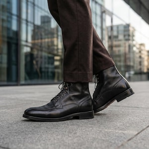 Peut inclure: Une paire de bottines à lacets en cuir noir avec un bout arrondi, des coutures visibles le long de l'empeigne et un petit talon. Les bottines sont portées avec un pantalon marron foncé, révélant la silhouette montante et les lacets noirs.