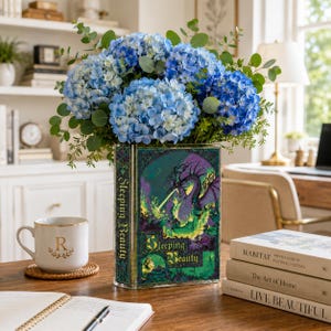 Op de afbeelding: Een rechthoekige glazen vaas met een Doornroosje boekomslagontwerp en een paarse drakenillustratie. De vaas is gevuld met een groot boeket blauwe hortensia's en groene eucalyptusstengels en staat op een houten bureauoppervlak.