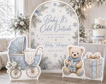 May include: A set of three decorative party standees featuring a large arched sign with the text Baby It's Cold Outside, A Winter Wonderland Baby Shower, Baby Harper, January 18, 2027, a blue baby carriage, a teddy bear, and a gift box.
