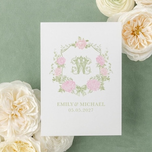 May include: A white invitation card featuring a floral wreath with pink roses and a green bow. The card includes the names "EMILY & MICHAEL" and the date "05.05.2027" in green. White and cream-colored flowers surround the card on a green background.
