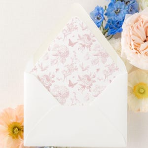 May include: A white envelope with a pink floral and butterfly pattern lining, surrounded by an assortment of fresh flowers in shades of white, peach, and blue. The envelope is open, showcasing the delicate design.