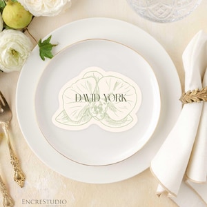 Puede incluir: Un cubierto con un plato blanco, un plato con borde dorado y una servilleta sujeta por un anillo dorado en forma de hoja. Una tarjeta de sitio con el nombre "DAVID YORK" está en el plato, con un diseño floral. Un tenedor y una cuchara dorados están a la izquierda.