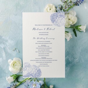 Pode incluir: Um cartão de programa de casamento branco com ilustrações de hortênsias azuis e texto azul escuro detalhando a ordem da cerimônia de Melissa e Robert em 25 de agosto de 2017 em Rivervale Barn, cercado por flores de ranúnculo branco e delphinium azul.
