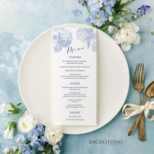 May include: A wedding menu card with a blue floral design. The menu includes starters, entree, and dessert options. The card is on a white plate with blue and white flowers.