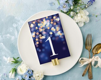 May include: A rectangular table number card featuring a dark blue background with a white number 1 and a bokeh light effect of gold and blue circles. The card is held by a small gold metal stand and placed on a white plate with blue flowers.