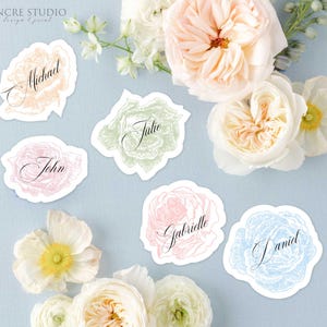 Puede incluir: Cinco tarjetas de sitio de papel troquelado con ilustraciones florales en melocotón, verde, rosa y azul. Cada tarjeta muestra un nombre en letra cursiva negra: Michael, John, Julie, Gabrielle y Daniel. El texto Encre Studio design & print es visible.