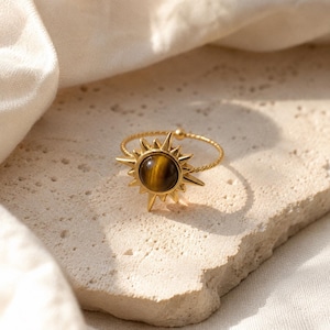 May include: A gold-toned ring featuring a central circular tiger's eye gemstone surrounded by a sunburst design with pointed rays. The band of the ring has a twisted rope texture, creating a detailed and cohesive piece of jewellery.