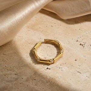 May include: A gold-toned open ring featuring a segmented bamboo-inspired design. The band is accented with four small, clear, round-cut stones embedded at intervals along the metallic surface, creating a continuous, textured, circular appearance.
