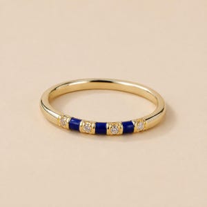 Puede incluir: Un anillo de banda dorada que presenta una serie de segmentos alternos de esmalte azul profundo y pequeñas piedras transparentes de corte redondo engastadas en el metal. El anillo tiene un acabado pulido y un perfil circular delgado y refinado.