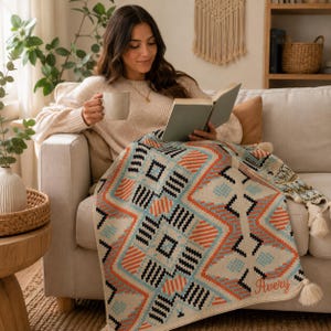 Peut inclure: Un plaid tissé présentant un motif géométrique dans des tons orange, bleu clair et noir sur fond crème, agrémenté de pompons crème aux coins. Le prénom Avery est brodé en écriture orange dans le coin inférieur de la couverture.