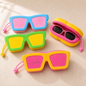 Peut inclure: Quatre étuis en forme de lunettes avec fermetures éclair et dragonnes. Un étui est bleu avec des verres roses, un est vert avec des verres jaunes, un est jaune avec des verres roses, et un est jaune avec des verres roses portant le nom Sophia.