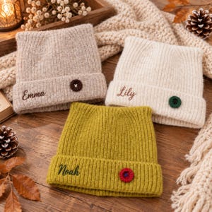 Peut inclure: Trois bonnets en tricot côtelé avec revers. Un bonnet beige brodé du prénom Emma en marron avec un bouton marron. Un bonnet crème brodé du prénom Lily en marron avec un bouton vert. Un bonnet vert olive brodé du prénom Noah en vert foncé avec un bouton rouge.