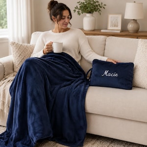Peut inclure: Une couverture jeté bleu marine en peluche à la texture douce, accompagnée d'une taie d'oreiller rectangulaire assortie avec le prénom Macie brodé en écriture cursive blanche. La couverture est drapée sur un canapé et la taie possède une poignée.