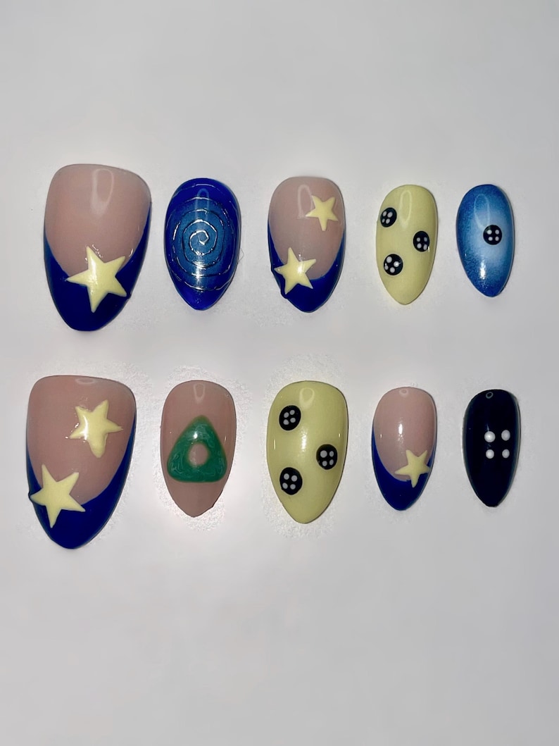 Puede incluir: Un juego de diez u&ntilde;as postizas con varios dise&ntilde;os, incluyendo puntas francesas azules con estrellas amarillo p&aacute;lido, una espiral azul, un tri&aacute;ngulo verde, u&ntilde;as amarillo p&aacute;lido con motivos de botones negros y u&ntilde;as azul oscuro con puntos.