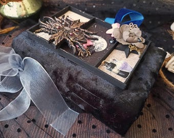 May include: A black velvet-covered box featuring a decorative lid with a large bronze-toned spider ornament, a beige flower, a crescent moon charm, and small paper cutouts of potion bottles, accented with a sheer light gray organza ribbon.