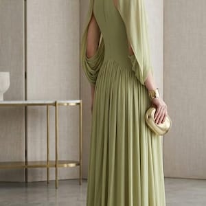 Puede incluir: Un vestido largo de color verde salvia con cuello alto, cuerpo ajustado y mangas largas y fluidas con aberturas en los hombros. La falda es plisada y llega hasta el suelo. Se incluye un brazalete dorado y un peque&ntilde;o bolso de mano dorado.